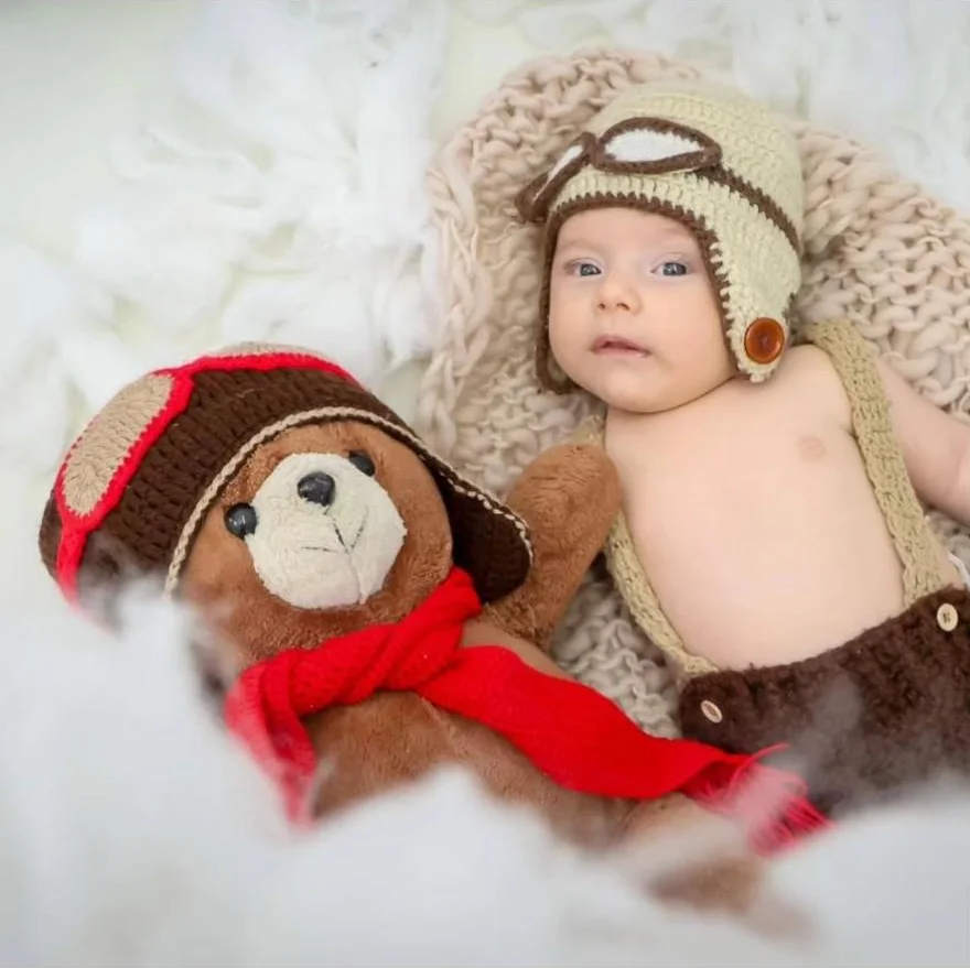 Bebê de gorro de aviador ao lado de um ursinho de pelúcia